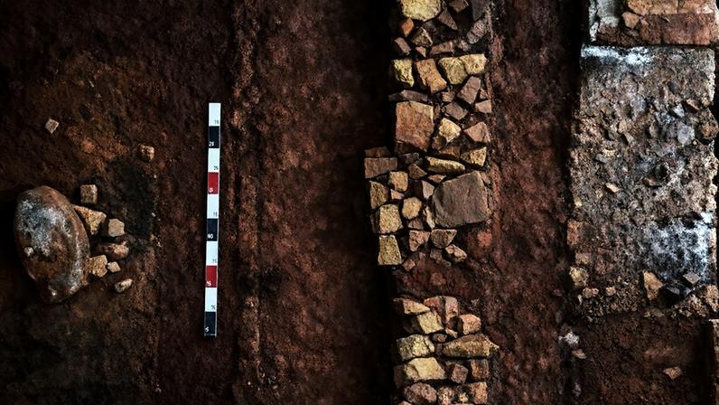 Chão de terra batida, de cor marrom, da antiga senzala da Fazenda do Pinhal. Na imagem, há manchas pretas e brancas, provenientes das fogueiras feitas pelos escravizados. No centro, observa-se os resquícios da estrutura da fundação de uma das paredes da senzala.