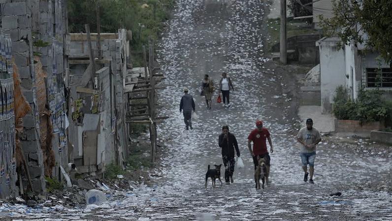Uma ladeira de terra. Pessoas com roupas de diferentes cores e dois cachorros, um preto e outro marrom, caminham pelo local aberto. O chão está repleto de papéis, sacolas e ao fundo um pouco de mato. Há algumas casas e muros nas laterais.