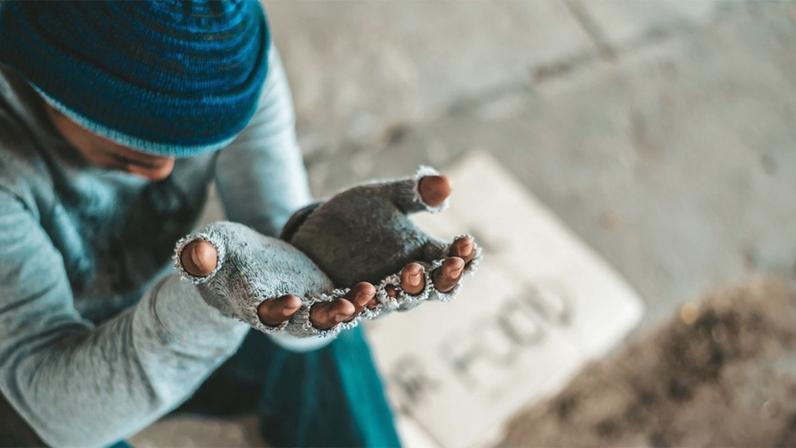Uma pessoa está sentada no chão usando um gorro azul e roupas em tons escuros. As mãos da pessoa estão estendidas para frente, abertas, como se estivesse pedindo algo. Ao lado dela, aparece um pedaço de papelão com escrita feita à mão, sugerindo um pedido de ajuda.