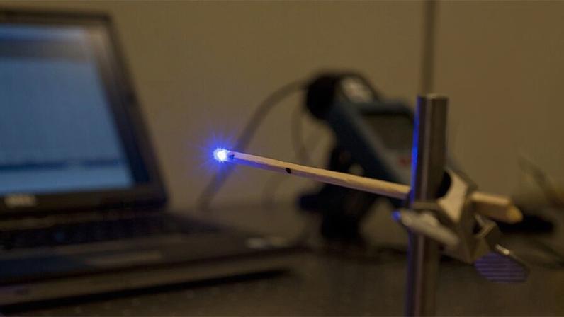 Equipamento de laser com o foco luminoso na ponta de um cabo preso a um suporte metálico, tendo ao fundo computadores e equipamento de medição
