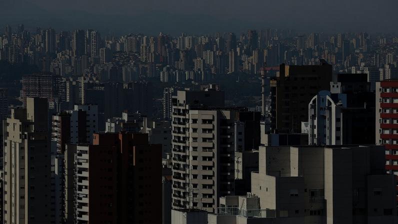 Vista da cidade de São Paulo com a cobertura de um edifício em primeiro plano, um conjunto de prédios com fachadas vermelhas, brancas, beges e azuis ao centro, e ao fundo, uma área verde e uma sequência de prédios.
