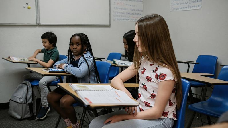 Sala de aula com crianças sentadas em carteiras azuis, lendo e escrevendo. Uma menina com tranças olha para a câmera enquanto as outras estão concentradas nos materiais escolares.