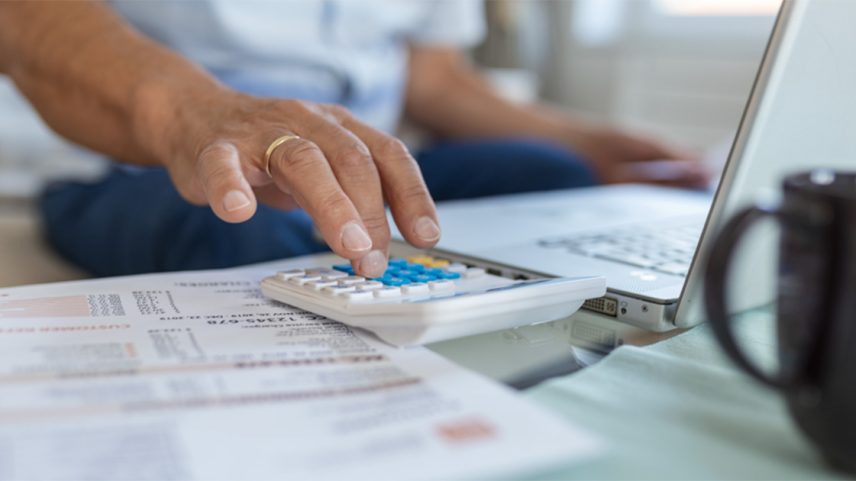 Com um notebook e uma fatura de contas a pagar na frente, uma pessoa calcula com ajuda de uma calculadora de mesa