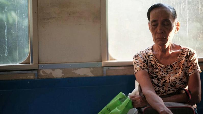 mulher de cabelos escuros e presos, sentada em um banco de trem segurando uma secola e com os olhos fechados, com aspecto de cansada