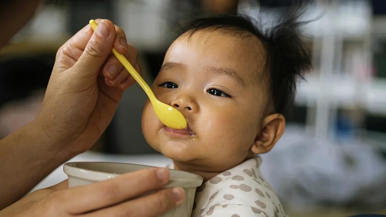 Mão adulta alimentando um bebê asiático com uma colher amarela, o bebê usa um macacão claro com bolinhas e tem um pouco de comida na boca