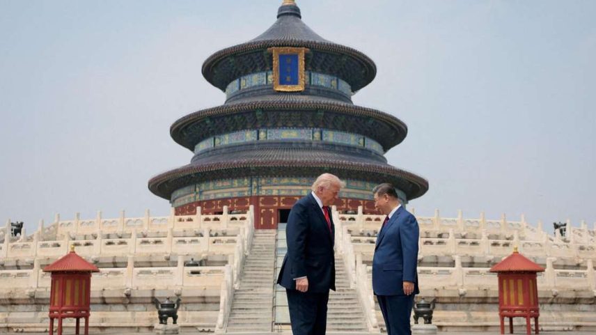 Os presidentes Trump e Xi estão em pé, frente a frente e com a cabeça levemente inclinada, diante de uma escadaria ornamentada que leva a um templo chinês decorado em tons de azul, verde, vermelho e dourado.