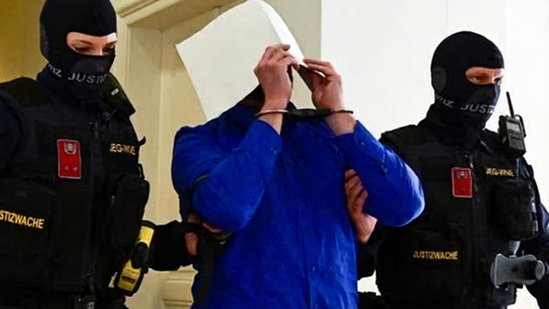 Beran A, covering his face with paper, being led along a corridor by prison guards