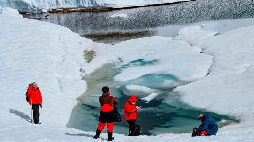 Quatro pessoas em roupas de inverno, três de casaco vermelho e uma de casaco azul, observam um lago de água azul-esverdeada cercado por neve e gelo. Uma pessoa à esquerda segura um bastão, duas ao centro olham para a água e uma à direita está agachada. Ao fundo, uma grande parede de gelo e um lago maior.