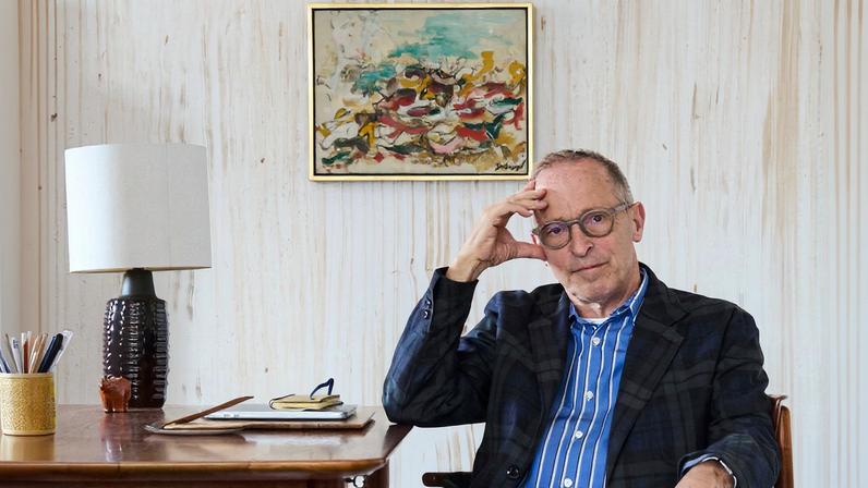 David Sedaris, wearing a blue-and-white striped shirt, a blazer and white pants sits at a desk in a room that is mostly empty aside from the desk and a bookshelf.