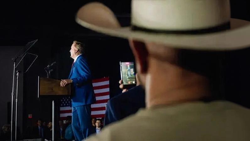 Ken Paxton, wearing a blue suit, stands at a lectern, with a large American flag hanging to his right.