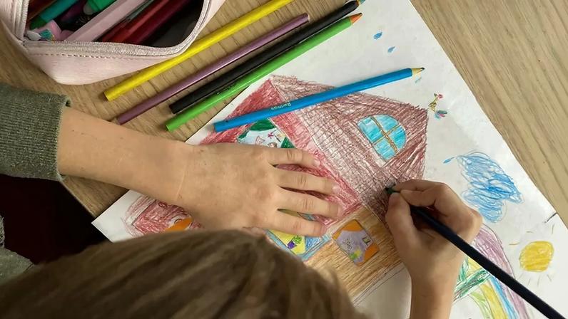 Criança com blusa verde desenhando uma casa vermelha e marrom em uma folha branca, usando lápis de cor verde. Lápis coloridos e um estojo rosa estão na mesa de madeira ao lado do desenho