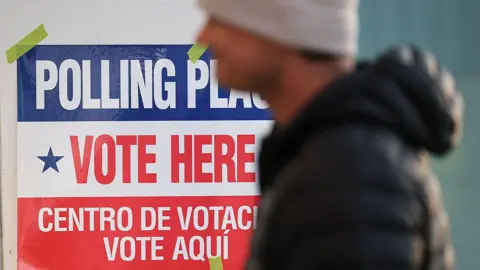 Getty Images A man in a beige tuque and a black puffer jacket walks by a red, white and blue sign that reads in English and in Spanish 'Polling place, Vote here".