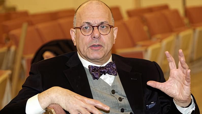 Leon Botstein in glasses, bow tie and vest gestures while speaking, holding a book