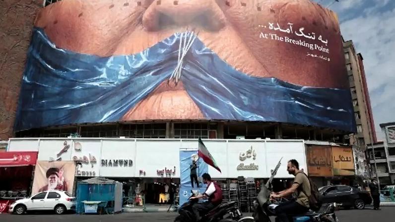 Motoristas passam diante de um painel anti‑EUA que faz referência ao presidente Donald Trump e ao estreito de Ormuz, instalado na praça Valiasr, em Teerã, em 2 de maio de 2026.
