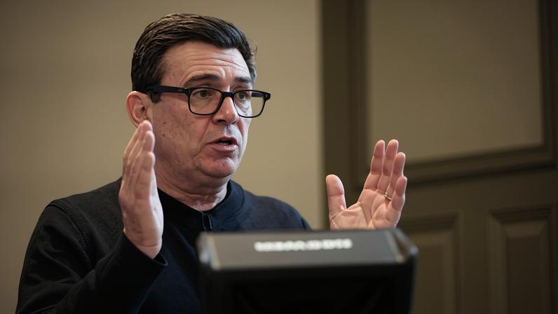 Andy Burnham speaks at an event. He is wearing a black jumper and black-rimmed glasses and is holding both hands up as he gesticulates.