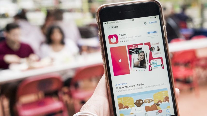 Pessoa usando celular mostra app Tinder na App Store, com fundo borrado de pessoas.