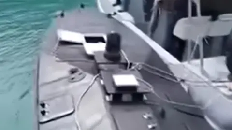 Footage has emerged showing the discovered naval drone (left) being towed by a Greek coastguard boat to the port of Vasiliki