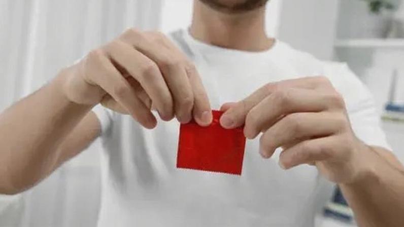 Getty Images A bearded man wearing a white t-shirt is seen opening a red condom packet with both of his hands inside a white coloured bedroom.