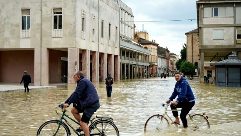 Dois homens pedalam bicicletas em rua de cidade alagada por água turva. Um carrossel está parcialmente imerso na água à esquerda.