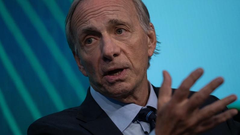 Ray Dalio, de terno e gravata, fala gesticulando com a mão aberta. Fundo azul.