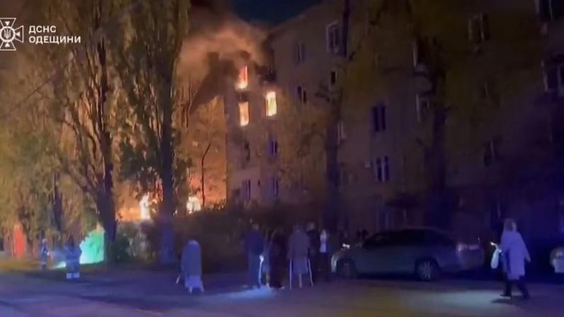Incêndio em prédio residencial na Ucrânia com pessoas observando de longe e bombeiros tentando controlar o fogo durante a noite