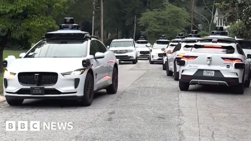 A litany of Waymo cars take up all the space in a street.