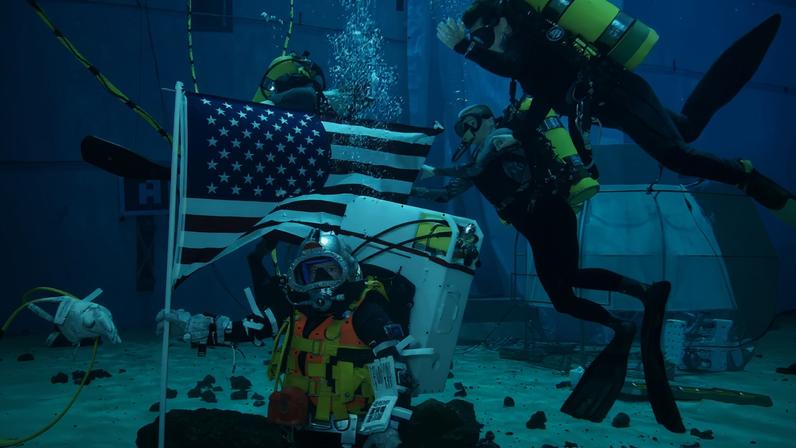 Two people practice underwater operations in a Moon like environment. The person on the left is holding an U.S. flag.