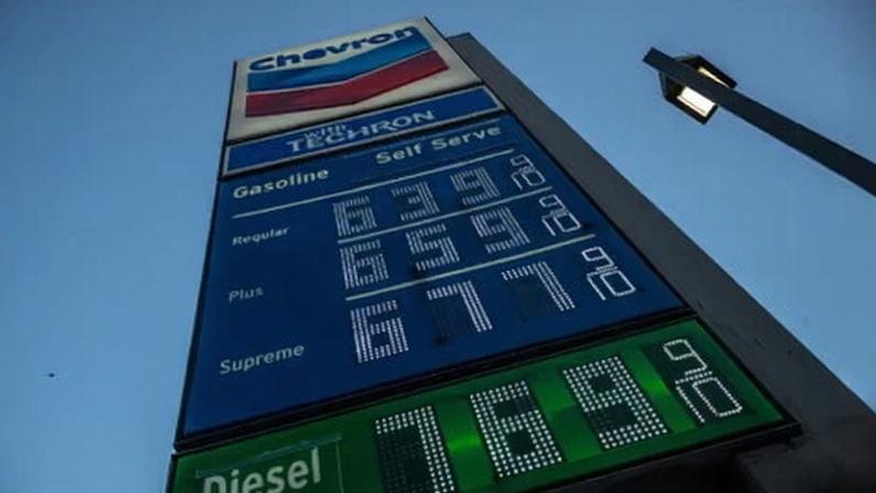 A Chevron gas station price sign displays fuel prices in LED digits against a blue sky