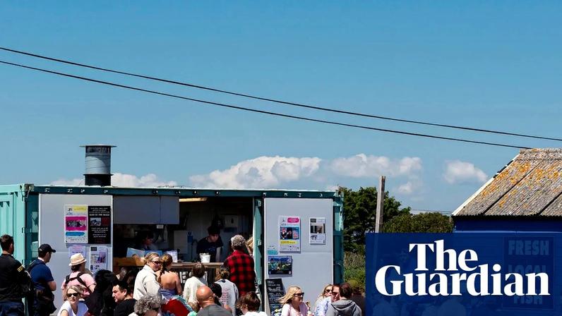 People sit at picnic benches outside a turquoise shipping container cafe and a blue shed with a 'Fresh Local Fish' sign.