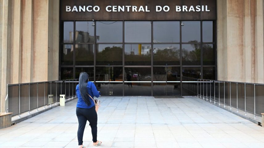 Pessoa de costas caminha em direção ao Banco Central do Brasil, prédio com fachada de vidro.