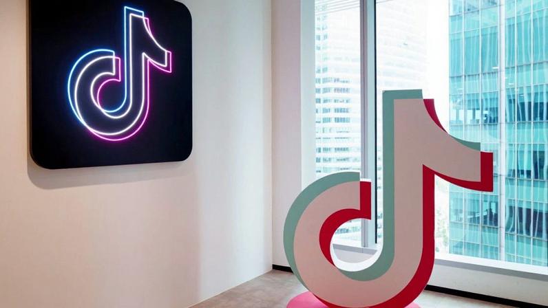 Dois logos do TikTok: um neon na parede e outro 3D no chão de um escritório moderno, com janela e prédios externos.
