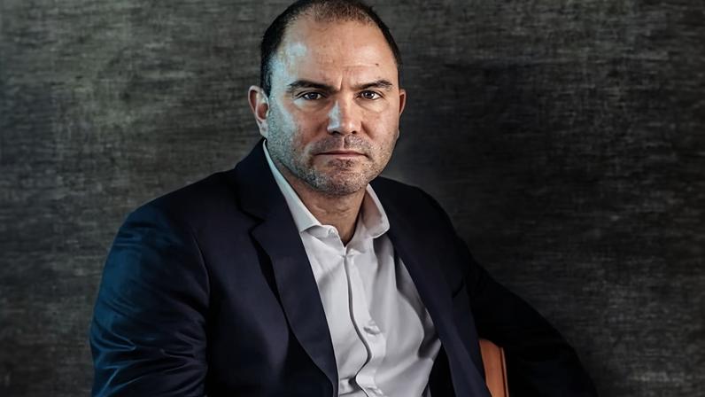 Ben Rhodes in a dark suit and white shirt sits at a table with hands clasped, looking directly at camera