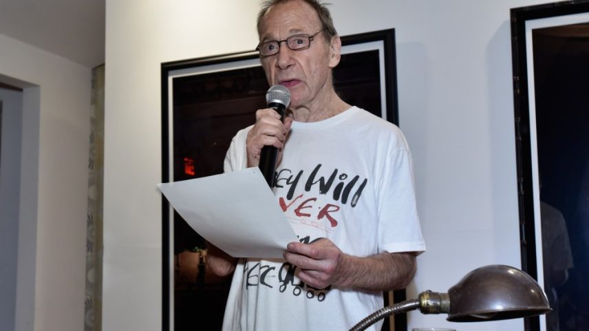 Anthony Haden-Guest, in a long white t-shirt and glasses, speaks in to a microphone.