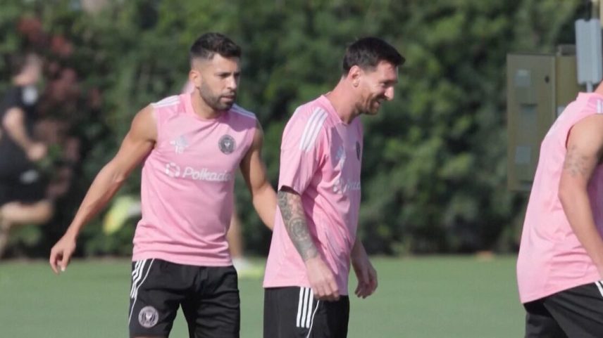 Lionel Messi e outro jogador treinando com uniforme rosa e shorts pretos em campo gramado.