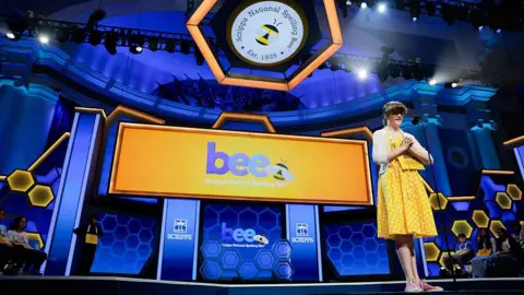 Getty Images A young girl in a yellow dress stands on a stage that is branded with National Spelling Bee logo