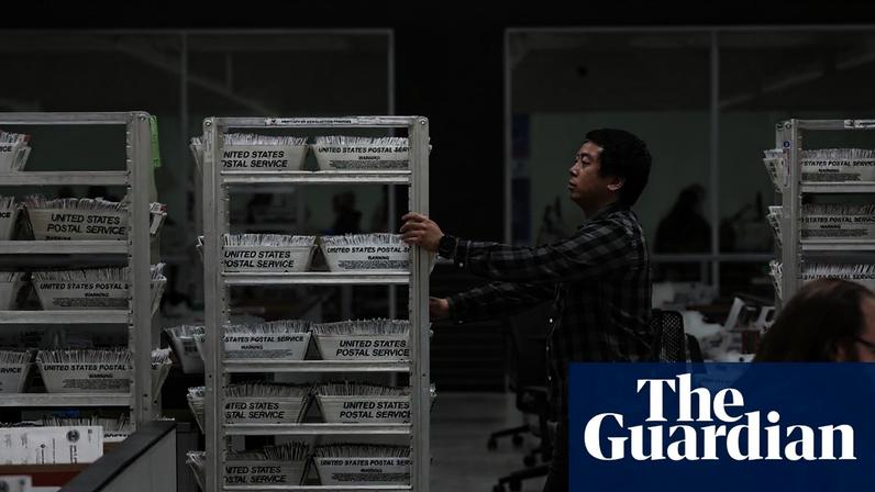 A worker handles trays of mail-in ballots labeled 'United States Postal Service' in a processing facility