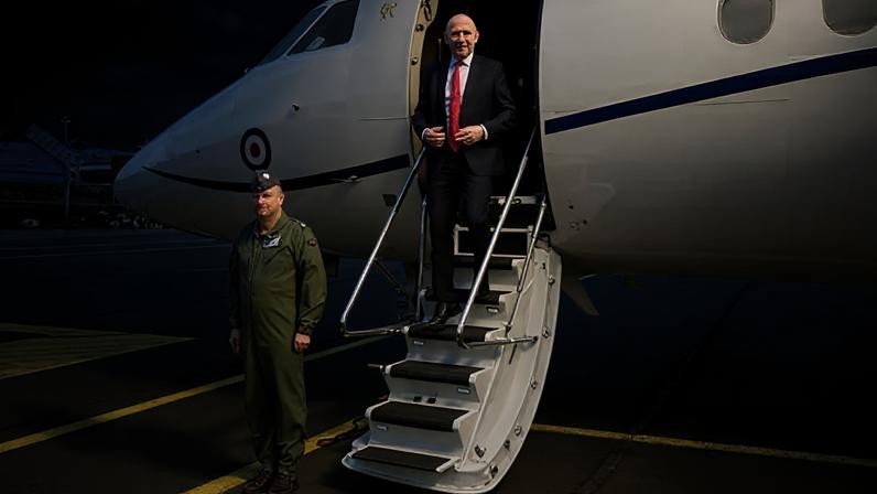 John Healey in a white shirt, black suit and red tie, seen exiting an RAF plane at an airport as a soldier stands to attention nearby