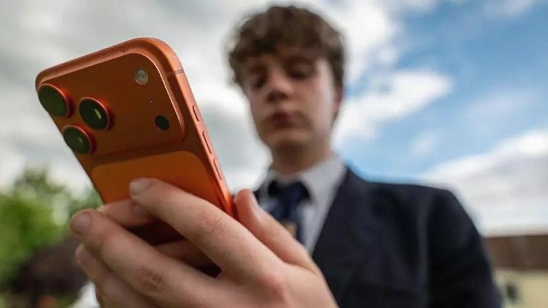 Um adolescente vestindo um uniforme escolar da Marinha, blazer, gravata e camisa branca segura um smartphone laranja à sua frente.