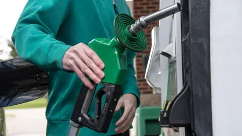 Adam J. Dewey/Anadolu via Getty Images A man in a green jumper returns a petrol pump after refilling his car