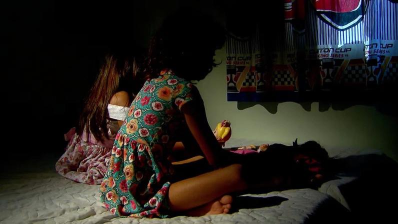 Duas crianças brincando com bonecas sentadas em uma cama em ambiente com pouca luz