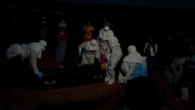 People in full PPE surround a coffin next to a grave, with other graves beside them
