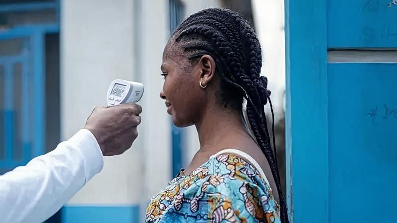 Getty Images A medic checks the temperature of a woman, using a contactless infrared thermometer. Only the arm of the medic, who is wearing white, is shown. The woman having her temperature checked has her hair in braids. She is wearing a bright blue top with a pattern and is standing next to a blue wooden door with a white and blue building in the background.