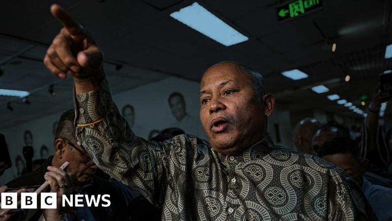 Philippine Senator Ronald Ronald Bato Dela Rosa gestures with his right hand, pointing upwards with his index finger while surrounded by journalists in the Senate hall
