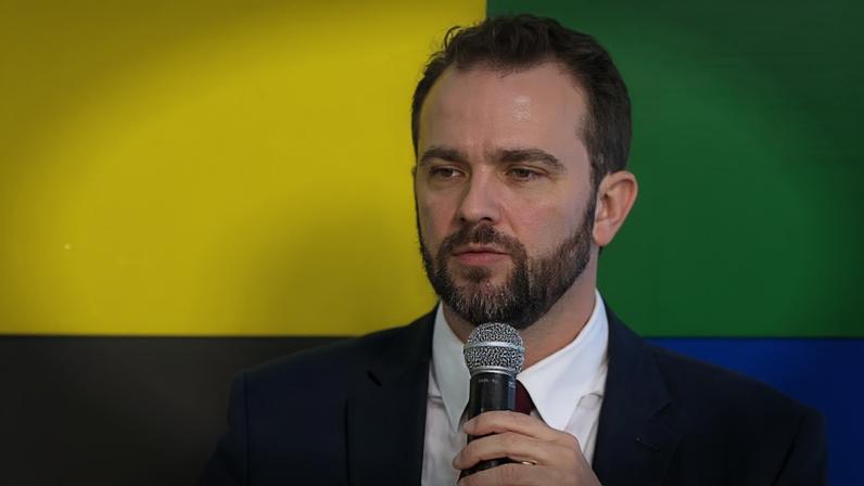 Homem de terno escuro, camisa branca e gravata vermelha fala ao microfone em evento oficial. Atrás dele, painel colorido com grandes blocos nas cores verde, amarelo, azul e branco.