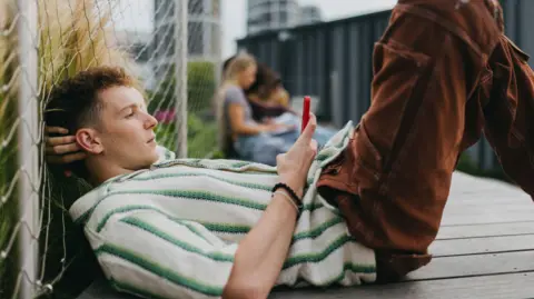 Getty Images A young man lying down on the ground holding and scrolling on his smartphone.