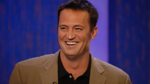 BBC US actor Matthew Perry laughs on set while wearing a black shirt and khaki blazer.