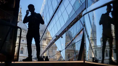 Getty Images A silhouetted man puts headphones in his ears, standing next to a modern glass building reflecting St Paul's cathedral in the background.