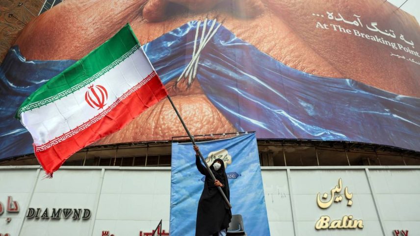 Uma mulher acena com uma bandeira iraniana em frente a um cartaz anti-EUA que faz referência ao presidente americano Donald Trump e ao Estreito de Ormuz, na Praça Valiasr, em Teerã, em 5 de maio de 2026.