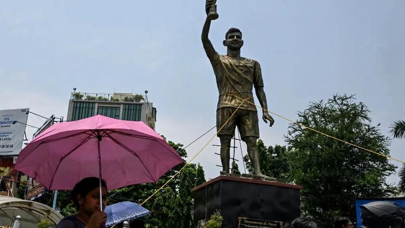 Pedestres caminham próximo à estátua de Lionel Messi em Calcutá, na Índia, em 27 de maio de 2026. Estrutura está presa por cordas devido à instabilidade e risco de queda