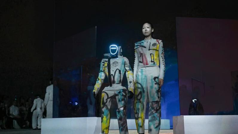 Uma modelo asiática e um robô humanoide, ambos vestindo macacões brancos com grafites coloridos, posam em uma passarela. O robô tem uma luz neon azul no rosto. O fundo é um telão com projeções de galáxias e estrelas em tons de vermelho, azul e amarelo.